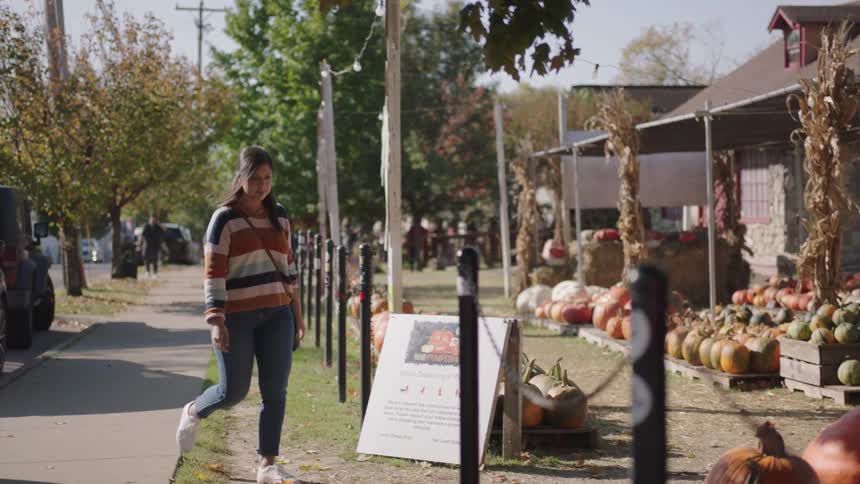 A young woman walks down a sidewalk and enters a produce stand to look at the pumpkins
