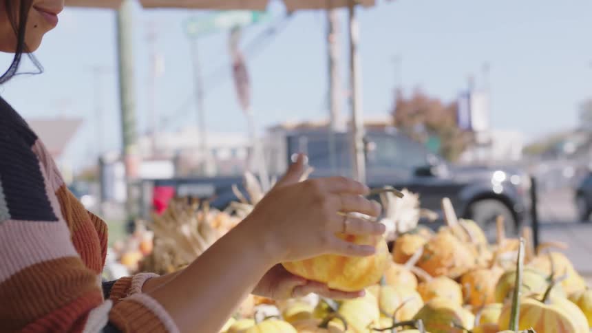 A woman picks up a squash to look at it and places it back in the box at an outdoor market on the corner of a street during the day