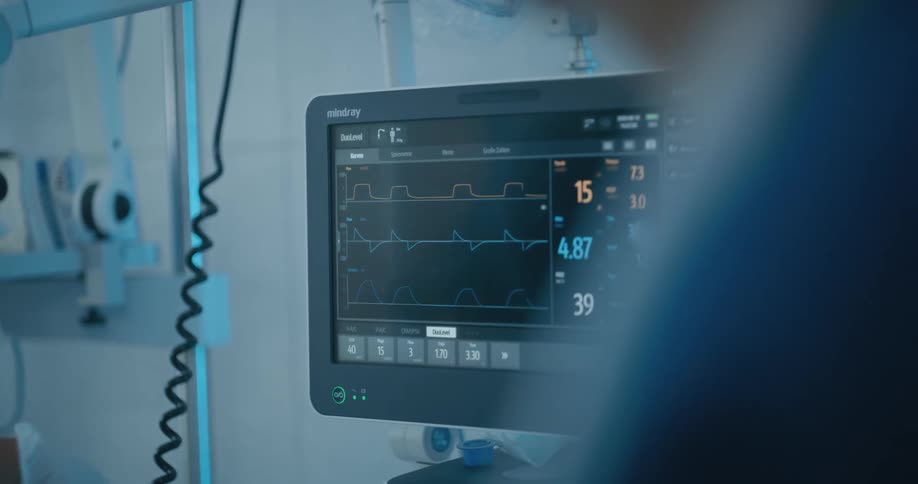 A pair of medical workers in personal protective equipment touch the screen of a vital signs monitor in a hospital