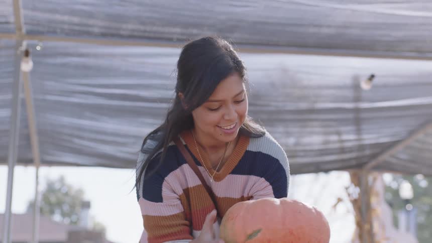 A woman bends down to pick up a pumpkin at an outdoor market on a busy corner of a street during the day