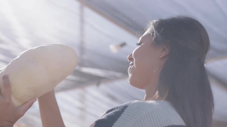 A young woman picks up a white pumpkin examines it and sets it down at a produce stand