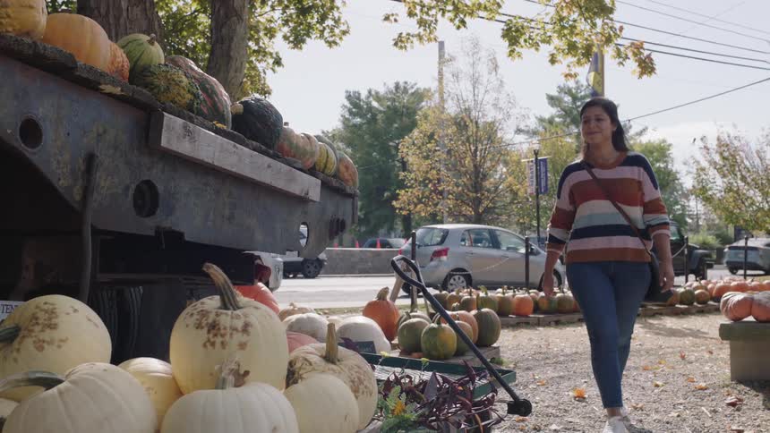 A woman walks into an open air farmers market and looks at a pumpkin