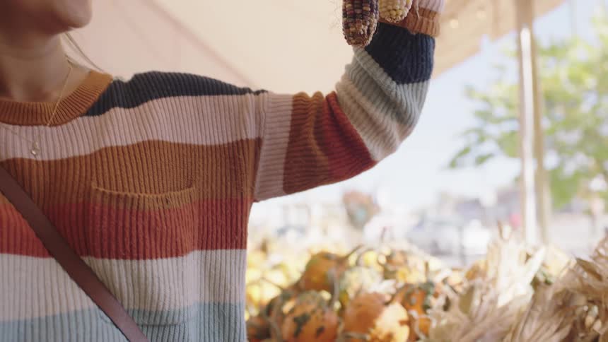 A young woman picks up a colorful bundle of indian corn and examines it at a produce stand