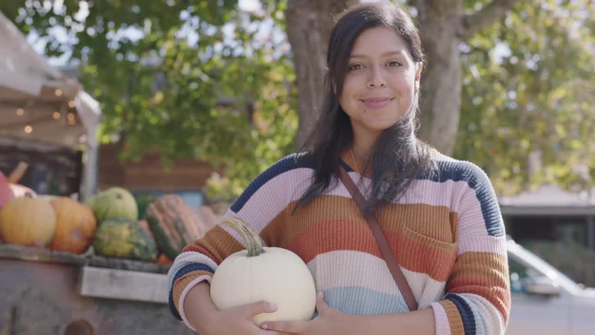 A young woman holding a white pumpkin smiles in front of a produce stand