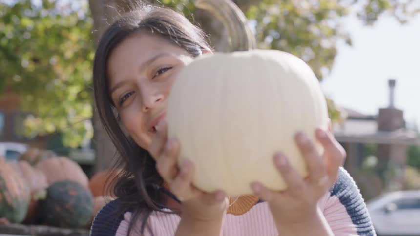 A teenage girl holds a pumpkin in front of her face and then peaks around it and smiles