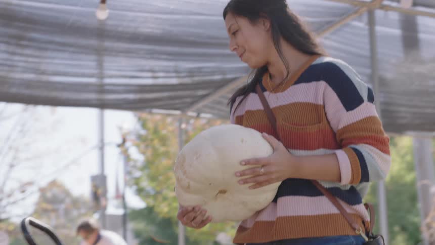 A young woman picks up a white pumpkin and places it in a wagon full of pumpkins at a produce stand