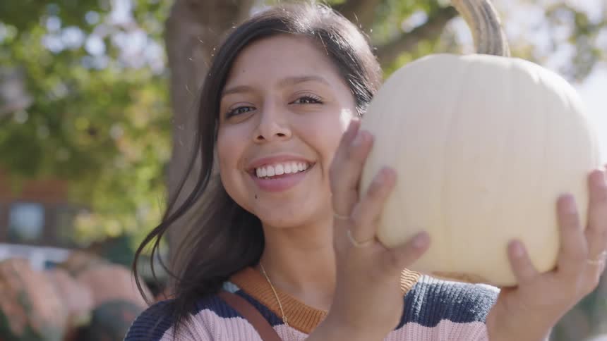 Brunette woman with dark straight hair holds up a white pumpkin and smiles at the camera in the market