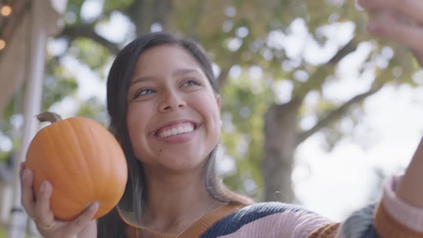 A teenage girl holding a pumpkin takes a selfie with her cell phone