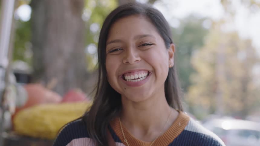 A woman holds up a pumpkin in front of her face and removes it to show herself smiling as she stands outside