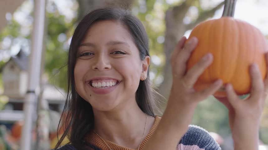 A teenager smiles as she pops her head out from behind a pumpkin she is holding