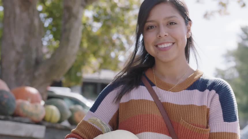 A woman in a striped sweater smiling while holding a pumpkin outside on a fall day