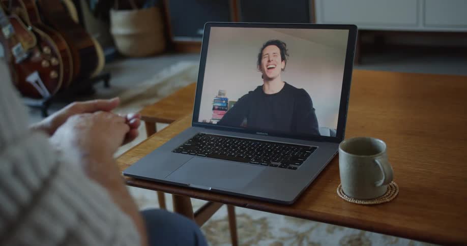 A man at home moves his hands around as he video chats with a friend who is lauging on a laptop