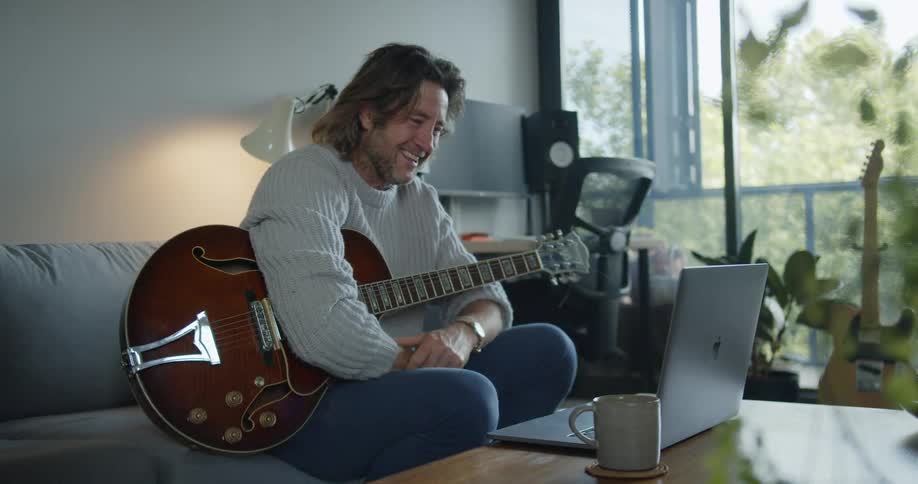 A middle aged man sitting on a sofa in a living room holds a guitar while participating in a video chat on his laptop