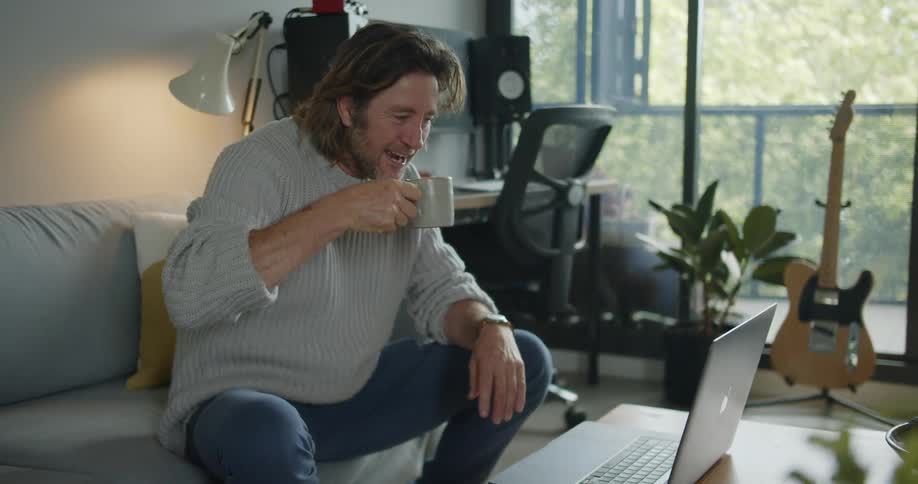 A man smiles and takes a sip of coffee while sitting on his couch during a video call on his laptop