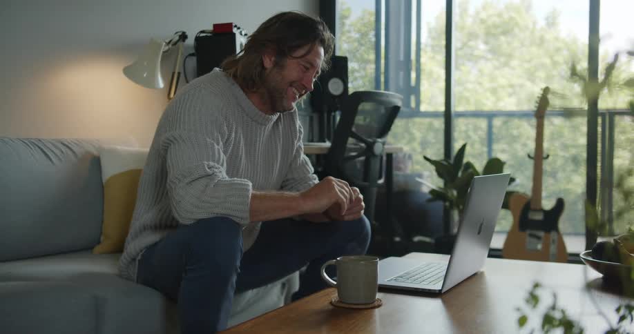 A man sits on a couch in a living room talking with a laptop open on a coffee table