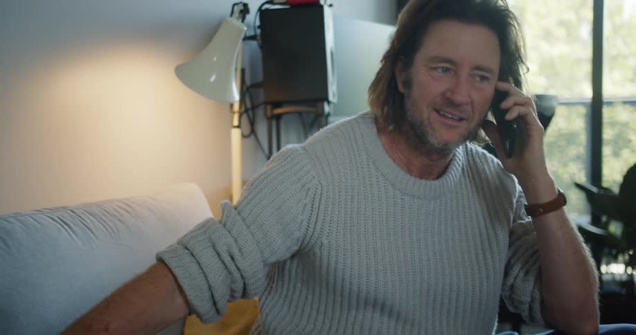 Man with a long hair and a grey pullover sits on sofa and pets brown dog while talking on smartphone in the living room at home