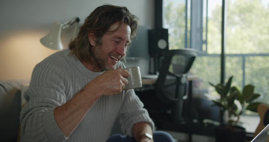A man participates in a video chat on his laptop while drinking from a steaming coffee mug in the living room of a house