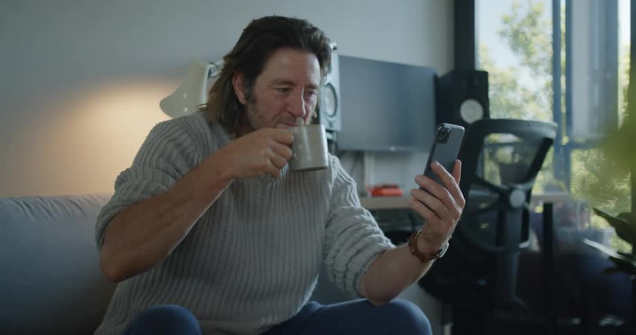 A man takes a drink of coffee while video chatting on a smart phone he is holding