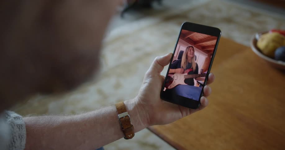 A man holds a cell phone with a woman displayed playing a guitar as he sits in a room at home