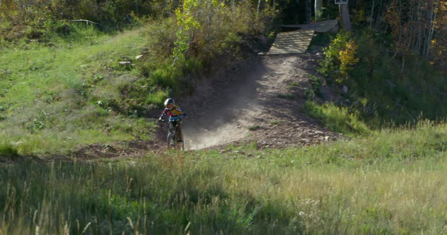 Two kids mountain bike down an incline in a forest