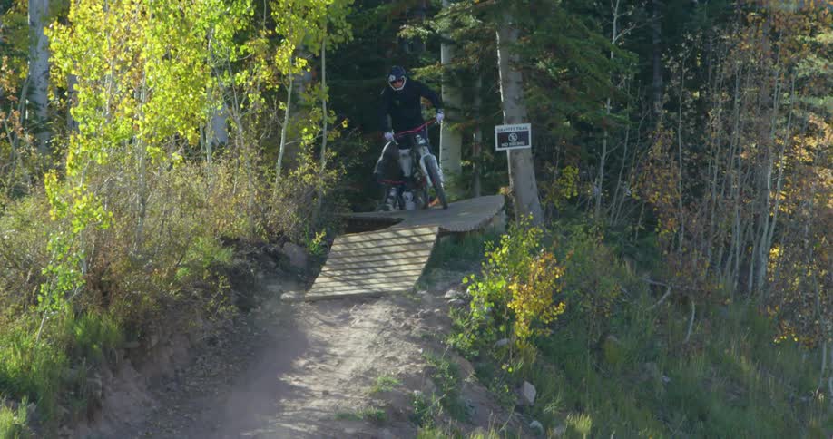 People on bikes exit a forest and ride down a hill on a dirt path during the day