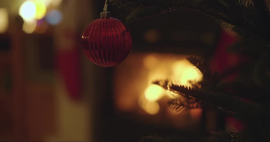 Red christmas ornament hanging from the branch of a pine tree in front of a fireplace