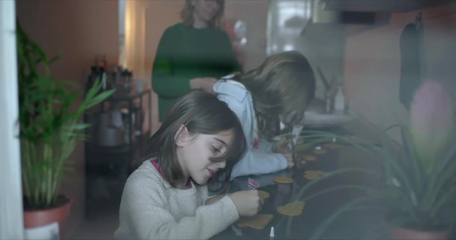 Two girls sit at a decorating decorating cookies that their mom places on the table