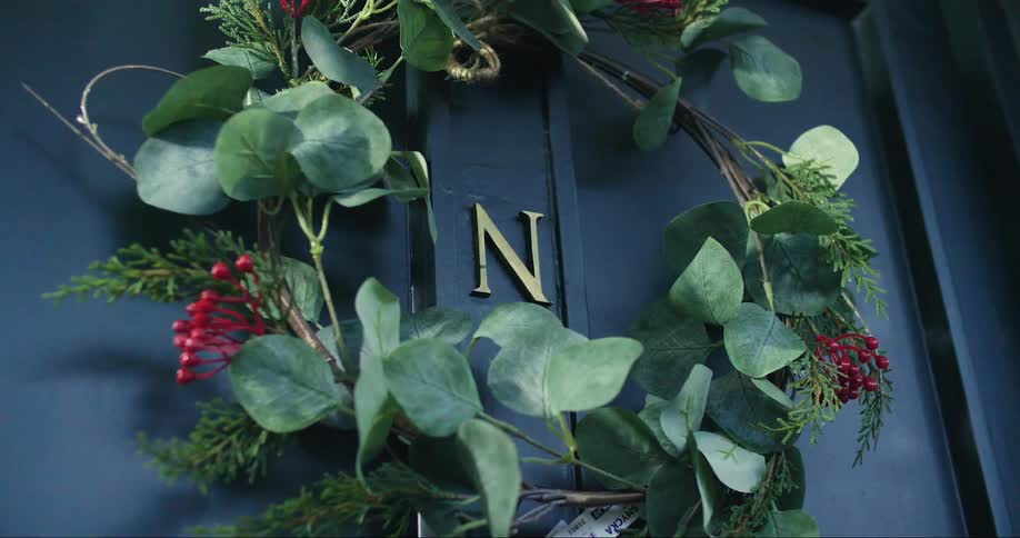 A wreath with red berries hangs on a blue door with a gold initial in the middle of the door