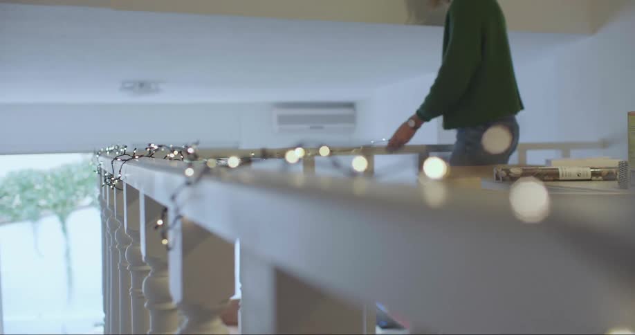 A woman stands on a second floor landing decorating a white railing with stringed lights