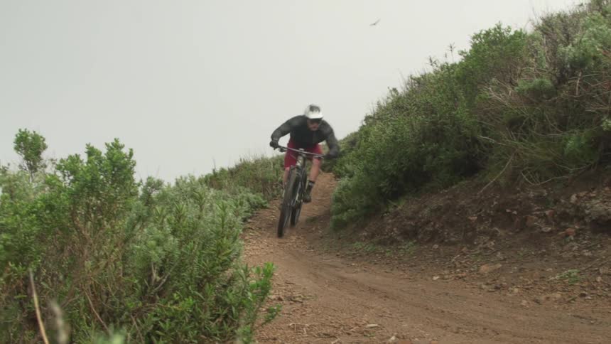 A cyclist rides a mountain bike along a narrow dirt path in the mountains at high speed on a cloudy day