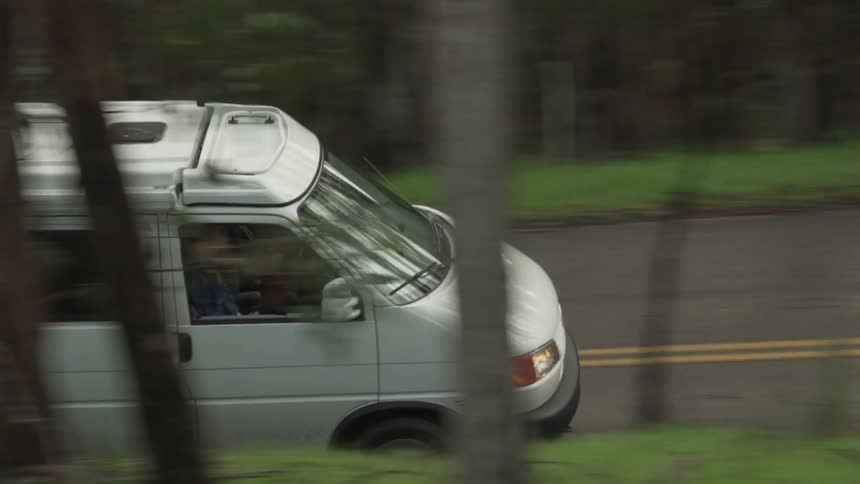 A white van drives down a rural two lane road that runs through a forest