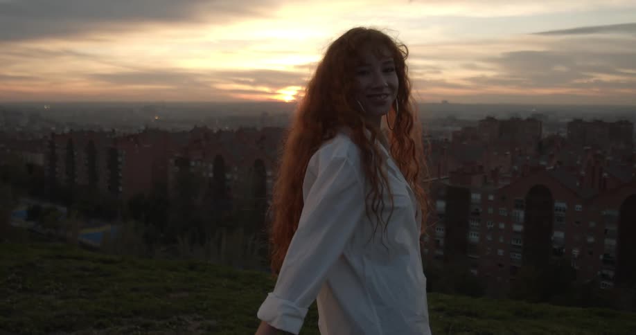 A woman with long red hair stands on the top of a hill overlooking building at dusk