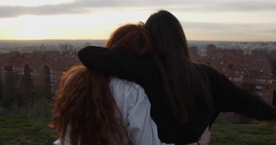 Two women embrace as they stand outside on a hill overlooking red brick buildings at sunset and raise their arms in the air