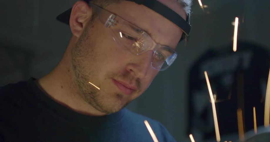 A man wears safety goggles as sparks fly close to his face while using welding equipment in his garage