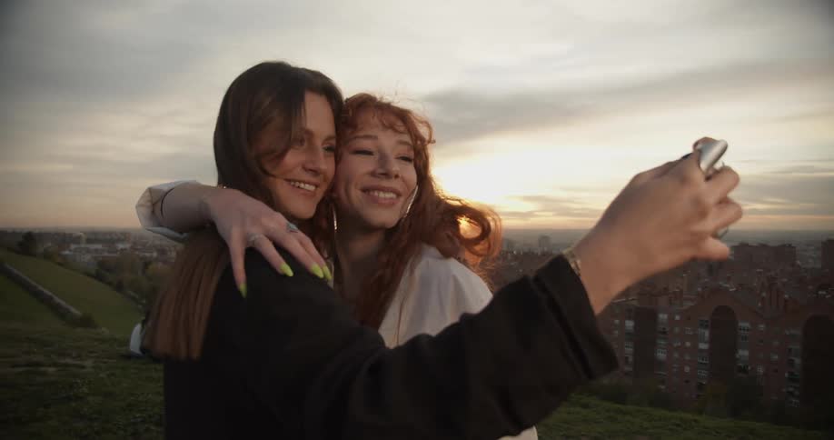 A girl holds her phone and takes a selfie as another girl kisses her