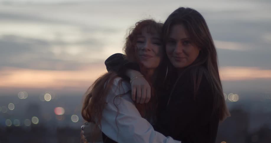 Two girl friends hugging and kissing each other looking at the camera at sunset