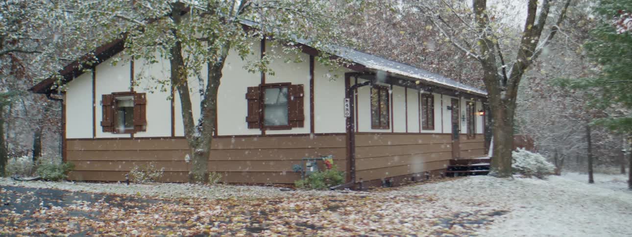 Snowflakes fall softly around a brown and white small home in a wooded area