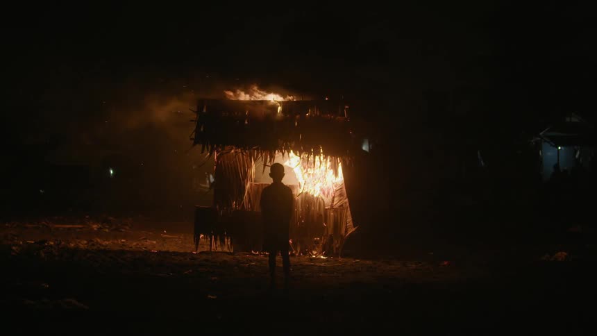 A boy stands in the dark in front of a burning structure