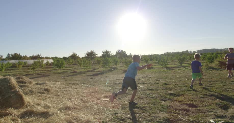 Three little boys outside farm backyard at daytime as one jumps off a stack of hay to run to the other two