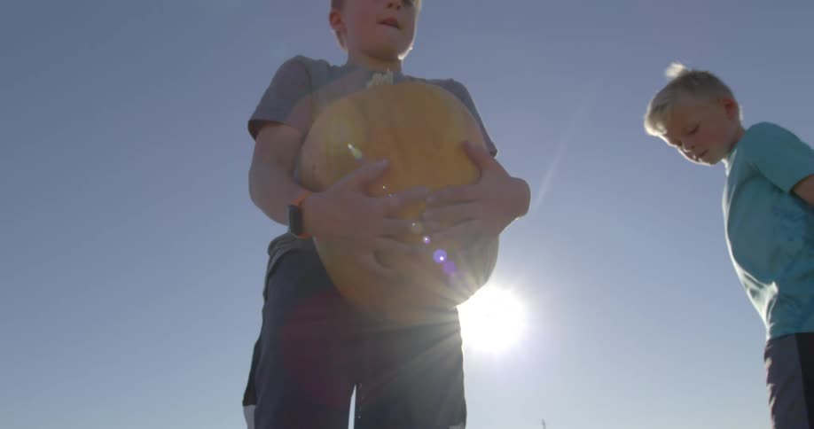 A white boy is picking up a big orange pumpkin in a pumpkin patch along side his brother who is searching for one under the blue sky with the sun beaming down
