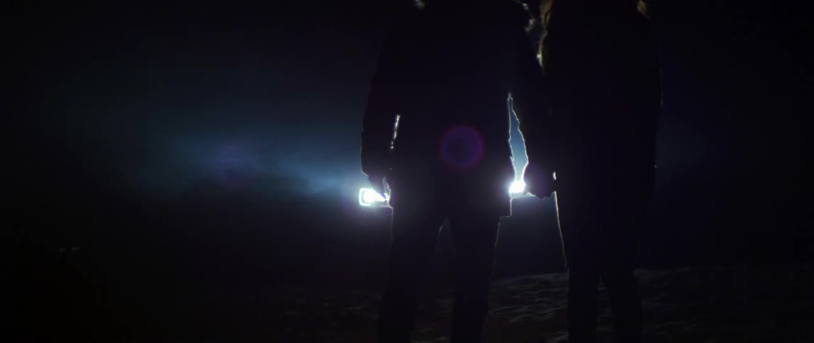 A silhouette of a couple standing in front of a parked car with its headlights on reach to hold each others hands