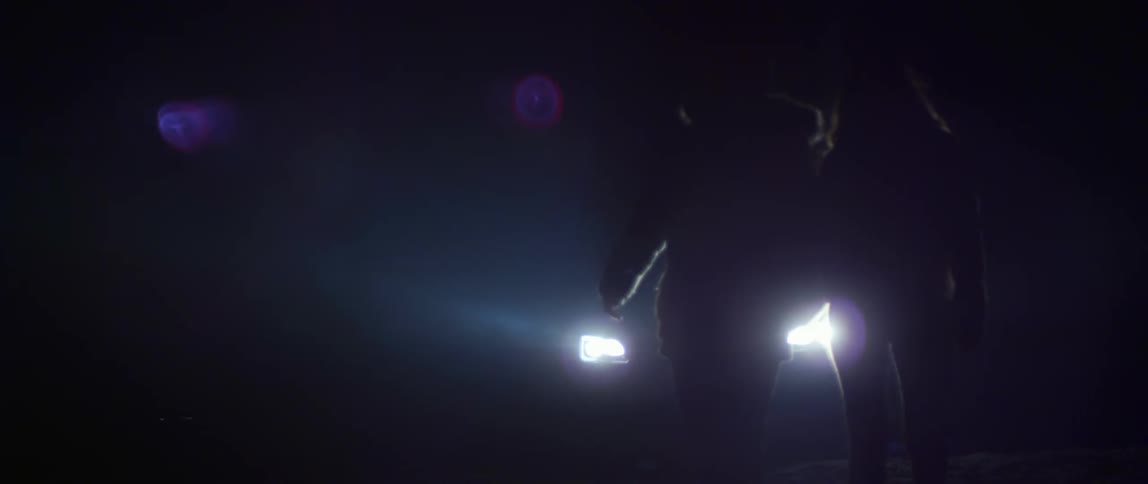 Two people holding hands walk in the dark in front of a vehicle with its lights on