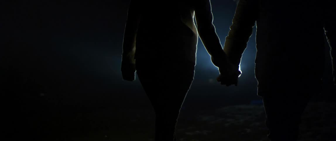 A man and woman reach for one anothers hands and hold hands as they walk towards a car with its lights on at night