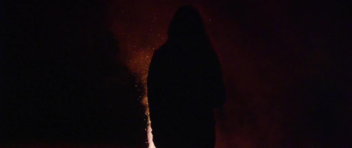 Silhouette of a hooded figure standing in front of fireworks at night