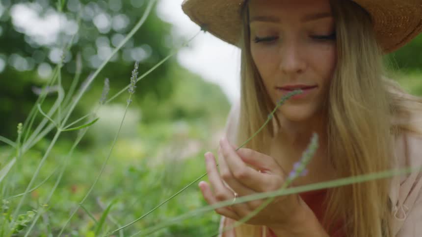 Woman wearing a sun hat sitting in a garden smells the flowers in the stem of a plant