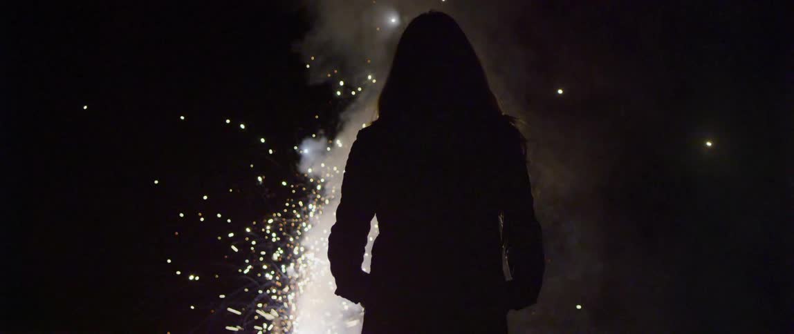 Silhouette of a robed figure standing in front of fireworks at night