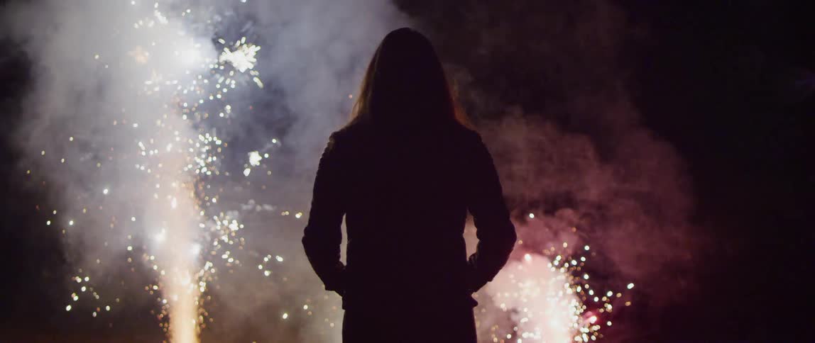 A man with long hair and wearing a jacket is enjoying a fireworks display at night