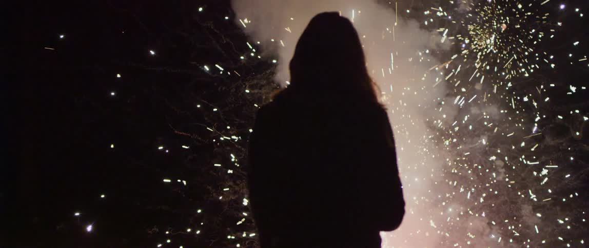 A woman stands outside watching fireworks in the sky at night