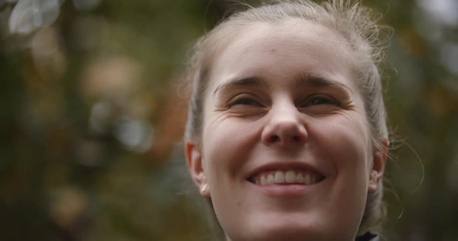 A young woman in a black jacket smiles and laughs while standing outdoors during the day