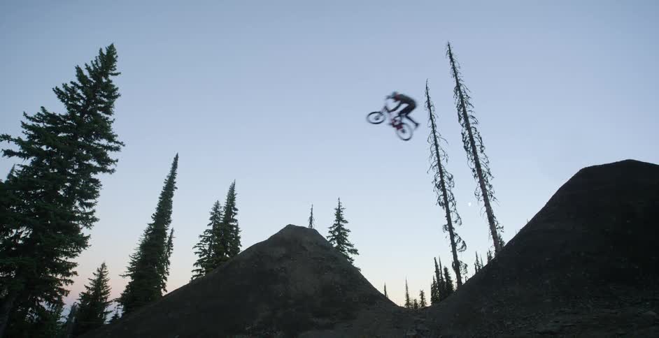 A cyclist flies through the air after riding a bicycle up a steep slope before landing on another slope in a woodland during the day
